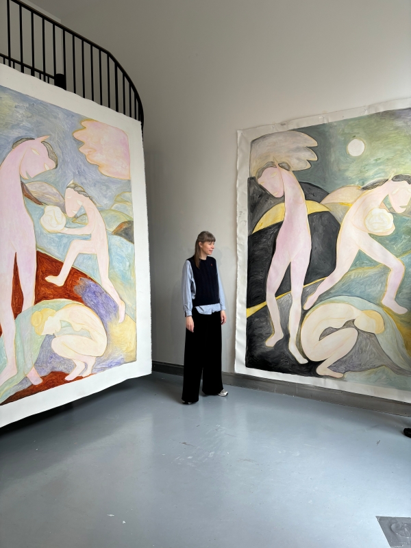 Die Künstlerin Sarah
                                          Bogner steht in einem hohen Ausstellungsraum zwischen zwei großformatigen Gemälden. Die Gemälde zeigen stilisierte, pastellfarbene
                                          Figuren mit angedeuteten Flügeln, fallweise mit Pferdeähnlichen Köpfen in traumartigen Landschaften. Die Künslterin lächelt.
                                          