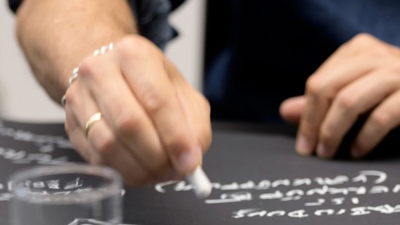 Foto einer Hand, die mit Kreide auf eine liegende Tafel schreibt.