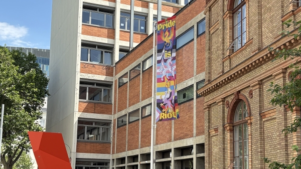 Eine Prideflagge vor der Universität für angewandte Kunst Wien. Darauf zu sehen ist ein Einhorn, ein Hippogreif
                                          und ein Phönix. Im Hintergrund gehen regenbogen-farbene Streifen ineinander über.