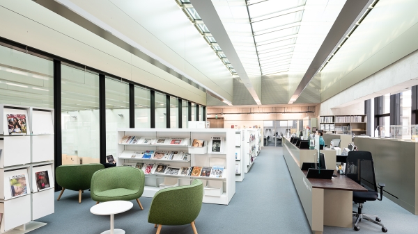 Interior view
                                          of the University Library at Vordere Zollamtsstraße 7: bright reading area with modern shelves, green armchairs, magazine
                                          displays, and the information desk under a glass ceiling.