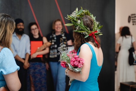 Foto einer Studierenden mit Blumenkranz und Zeugnis vor einer Gruppe Mitstudierender.